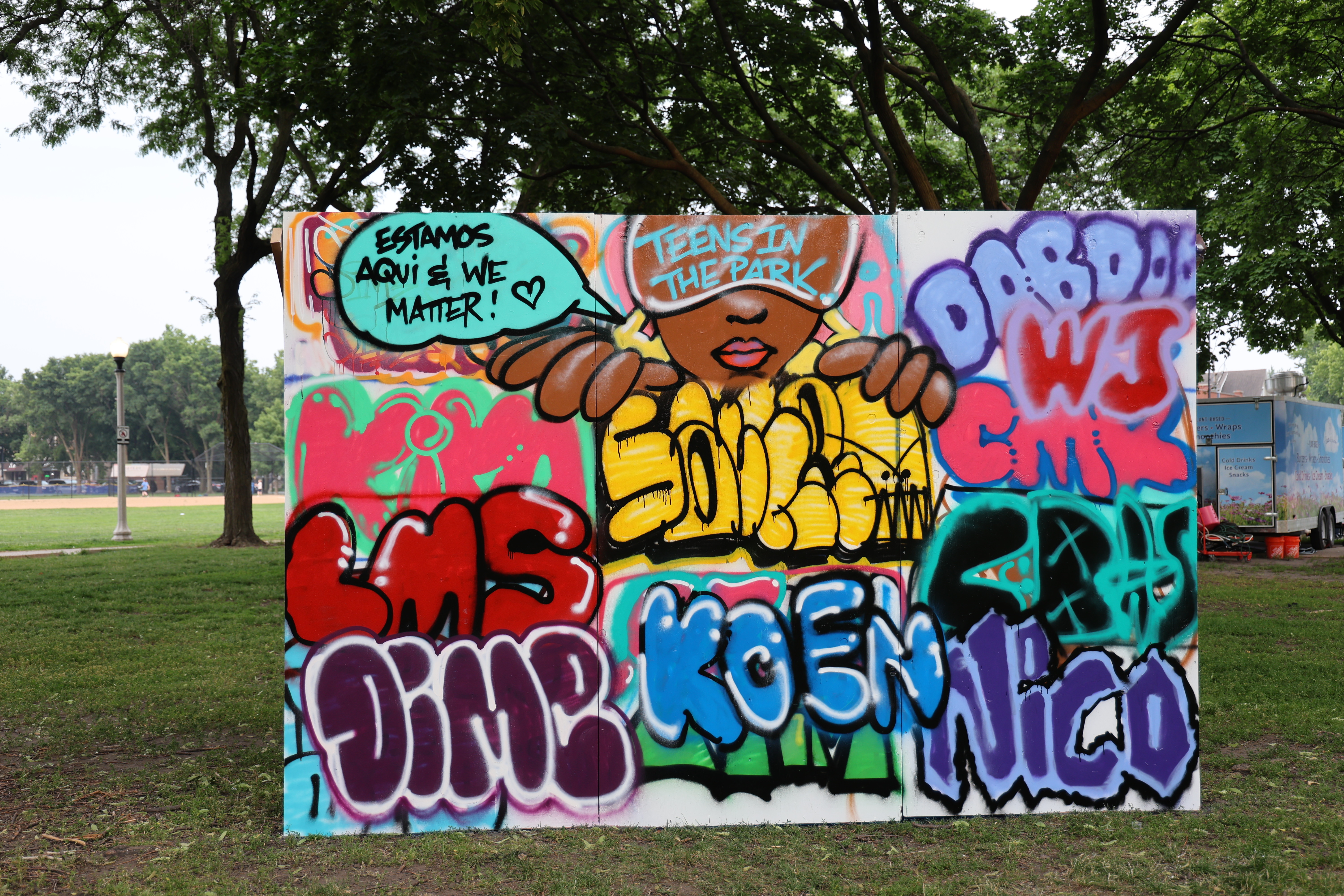 Colorful graffiti art on a large board in a park setting.  A face and hands are partially visible amongst various words and lettering. A speech bubble reads "Estamos Aqui & We Matter!" and another says "Teens in the Park".